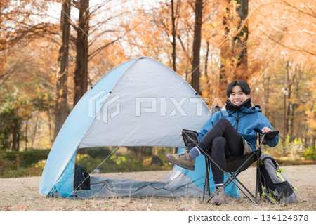 一位獨自露營的男士在秋季露營地的椅子上放鬆，享受戶外樂趣——他正看著鏡頭。 134124878