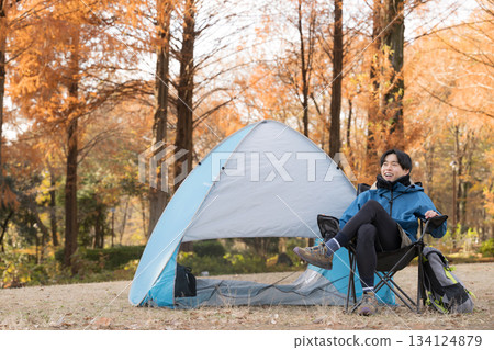 一位獨自露營的男士在秋季露營地的椅子上放鬆，享受戶外樂趣——他正看著鏡頭。 134124879