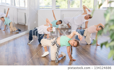 Smiling sports teen girl performing krump movements on floor with group children in breakdance studio with male coach 134124886