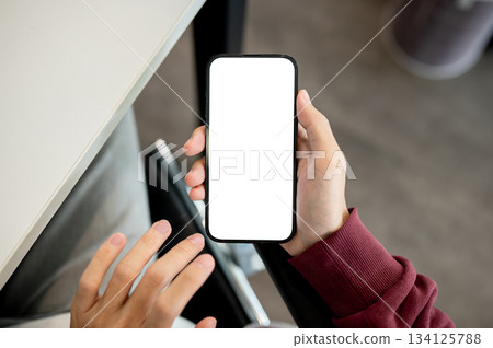 A man's hand holding white screen smartphone with fingers hovering aside at table in cafe or library 134125788