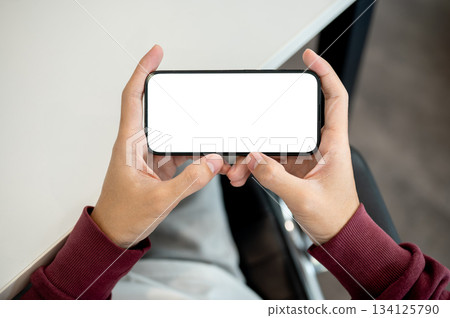 A man holding white screen smartphone horizontally as sitting indoors at table in a cafe or library. A man holding white screen smartphone horizontally as sitting indoors at table in a cafe or library. 134125790
