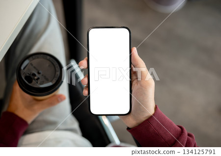 A man holding white screen smartphone aside coffee while sitting indoors at table in cafe or library A man holding white screen smartphone aside coffee while sitting indoors at table in cafe or library 134125791