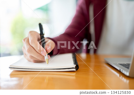 Male student's hand holding pen writing in notebook aside laptop on wooden table in cafe or library. 134125830