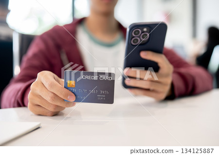 Close up man's hand holding phone looking at credit card sitting indoors at table in cafe or office Close up man's hand holding phone looking at credit card sitting indoors at table in cafe or office 134125887