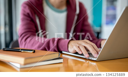 Close up man student's hand typing on laptop aside books and pen on wooden table in cafe or library. Close up man student's hand typing on laptop aside books and pen on wooden table in cafe or library. 134125889