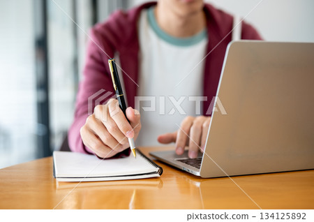A man student's hand holding pen over notebook typing on laptop at wooden table in cafe or library. A man student's hand holding pen over notebook typing on laptop at wooden table in cafe or library. 134125892