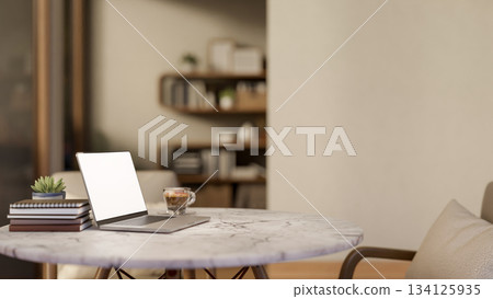 White screen laptop and coffee cup aside book on round marble table and wooden chair in living room. 134125935