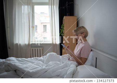 Happy middle aged woman procrastinating morning time in bed scrolling social media on smartphone. 134127373