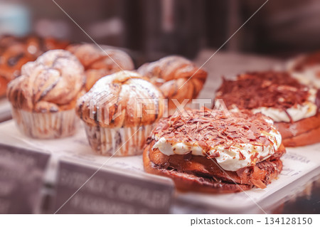 Fresh confectionery in the bakery window in close-up - brown muffins and muffins with powdered sugar and swirl dough, sweet rolls with chocolate cream and other desserts. High quality photo Fresh confectionery in the bakery window in close-up - brown muffins and muffins with powdered sugar and swirl dough, sweet rolls with chocolate cream and other desserts. High quality photo 134128150
