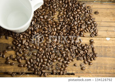 Flat design showing spilled medium-roast coffee beans and white ceramic mug on rustic wooden table Flat design showing spilled medium-roast coffee beans and white ceramic mug on rustic wooden table 134128792