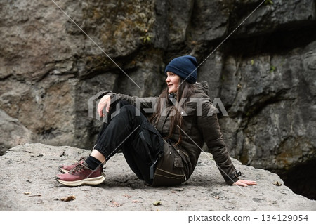 Smiling Russian woman resting on a rock during a hike near a mountain waterfall. Outdoor adventure and exploration in nature. Smiling Russian woman resting on a rock during a hike near a mountain waterfall. Outdoor adventure and exploration in nature. 134129054