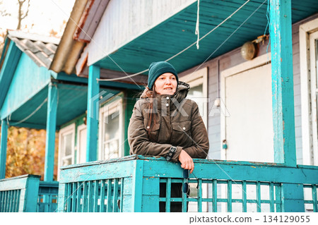 Russian woman standing on the porch of her rural wooden house, wearing warm clothes and hat. Cozy village life in autumn, winter or early spring. 134129055