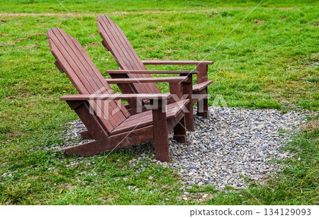 Two wooden Adirondack chairs on a small gravel area in a green grassy field, creating a peaceful outdoor seating background with copy space and nobody around. 134129093