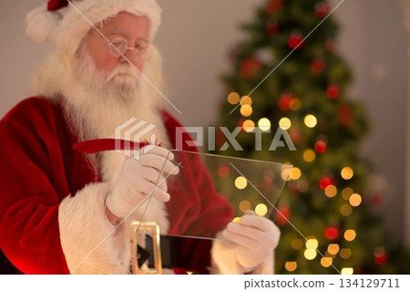 Santa Claus is writing on transparent tablet with red quill by lit tree in flat vector 134129711