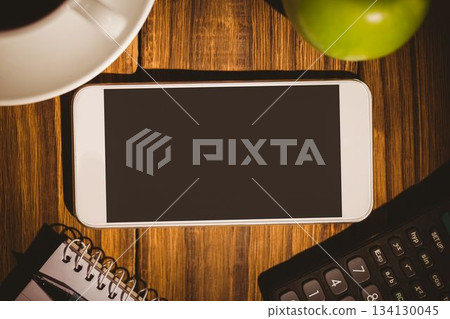 White-framed smartphone is lying on wooden desk with coffee cup, green apple, notebook, calculator 134130045