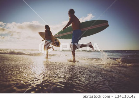 Low angle view of cheerful caucasian couple with surfboards running in sea against sky at sunset 134130142
