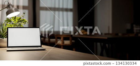 White screen laptop on wooden table under lamp light in dim lit living room or dining room. White screen laptop on wooden table under lamp light in dim lit living room or dining room. 134130334