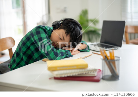 Asian kid elementary school boy wearing headphones sleeping over a book on table with laptop aside. Asian kid elementary school boy wearing headphones sleeping over a book on table with laptop aside. 134130606