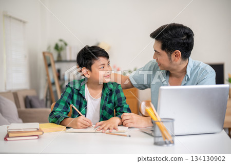 Father and a son smiling at each other while teaching and doing a homework together in their house. Father and a son smiling at each other while teaching and doing a homework together in their house. 134130902