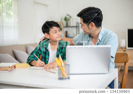 Father and son looking at each other while teaching and doing a homework together in the house. Father and son looking at each other while teaching and doing a homework together in the house. 134130903