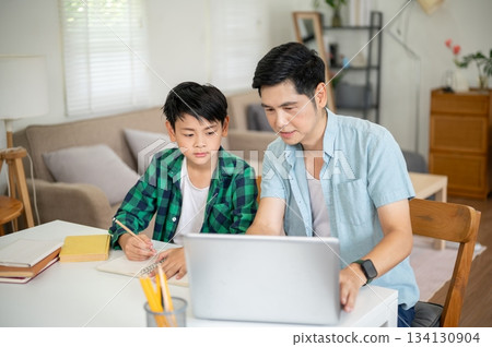 Father showing a laptop screen to his son while teaching and doing homework together in their house. 134130904