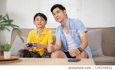 Asian father looking ahead with elementary school son holding joystick playing game at sofa in house 134130932