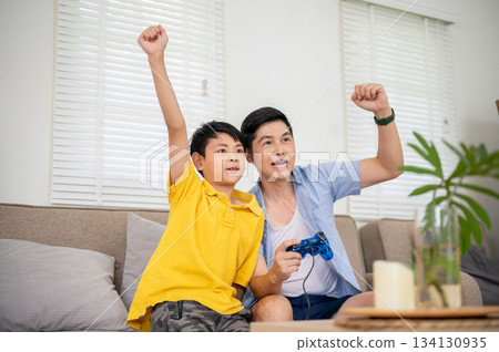 Asian father holding joystick playing game and raise a fist with his elementary son at sofa in house 134130935