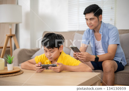 Asian father and his elementary son watching video on phone while sitting in their house living room 134130939