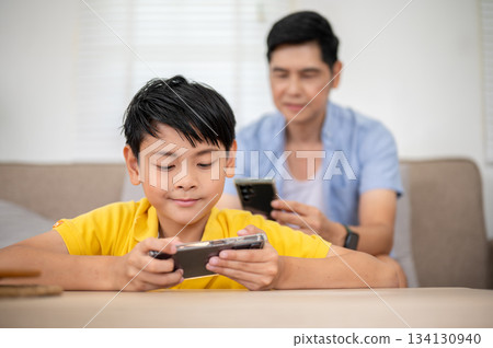 Asian father and his elementary school son using smartphone while sitting in their house living room 134130940