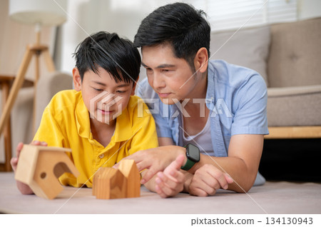 Close up of father and his elementary son looking at wooden house block toy on carpet in living room 134130943