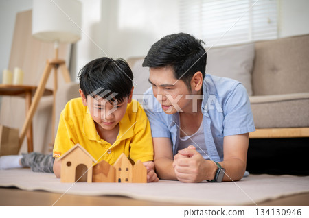 Close up of father looking at elementary son playing with wooden house toy on carpet in living room. 134130946