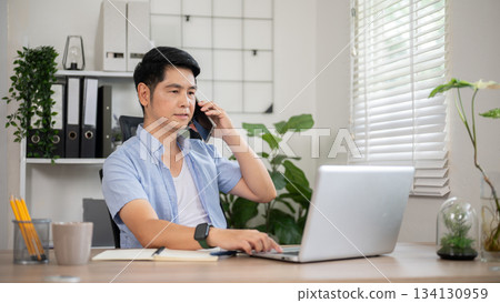 Asian man talking on phone and looking at laptop sitting at wooden table in office or working room. Asian man talking on phone and looking at laptop sitting at wooden table in office or working room. 134130959
