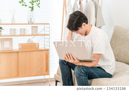 一位年輕的亞裔男子坐在沙發上使用筆記型電腦(正在調查,震驚,沮喪,犯錯) 一位年輕的亞裔男子坐在沙發上使用筆記型電腦(正在調查,震驚,沮喪,犯錯) 134133186