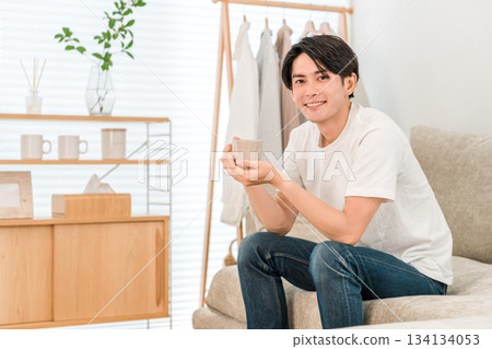 Young Asian man sitting on sofa in living room drinking hot drink, coffee, tea Young Asian man sitting on sofa in living room drinking hot drink, coffee, tea 134134053