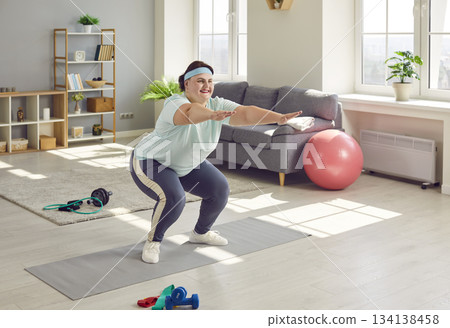 Happy young overweight woman doing squats during an active sports workout at home Happy young overweight woman doing squats during an active sports workout at home 134138458