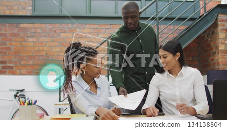 Reviewing printed documents, three coworkers gathering around desk in loft-style office with laptop 134138804