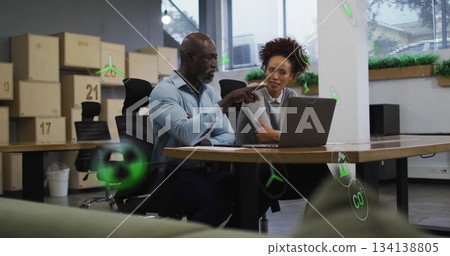 Collaborating colleagues presenting sustainability metrics on laptop in office, green icons 134138805