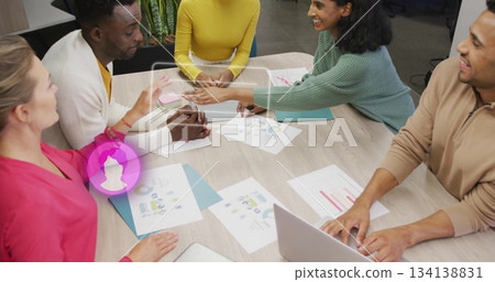Woman in mint sweater handing stylus across table in modern office, with printed charts and laptop 134138831