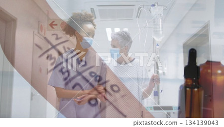 Walking mask-wearing nurse in scrubs guiding patient in gown with IV pole along hospital corridor 134139043