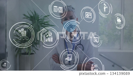 Standing senior female doctor in lab coat in hospital corridor, with stethoscope, digital icons 134139089