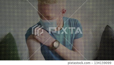 Man wearing protective face mask pulling up shirt sleeve and touching arm at home, with wristwatch Man wearing protective face mask pulling up shirt sleeve and touching arm at home, with wristwatch 134139099