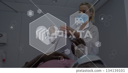 Masked doctor examining patient arm on exam table in exam room, with medical icons, copy space 134139101