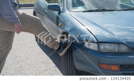 Caucasian man tries to fix damage to car body with his foot. Caucasian man tries to fix damage to car body with his foot. 134139405