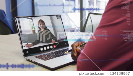Sitting man in burgundy shirt joining call on laptop in glass-walled office, with pen, notebook 134139967