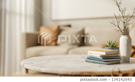 Books with flower vase and potted plant on round marble table across sofa in a blurred living room. 134142043