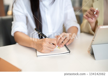 Close up of woman student's hand holding pen writing in notebook with teacher or supervisor talking. Close up of woman student's hand holding pen writing in notebook with teacher or supervisor talking. 134142082