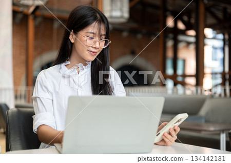 Asian woman student with glasses holding and looking at phone sitting with laptop at table in cafe. 134142181