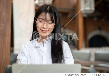 Asian woman student wearing eyeglasses looking at laptop while sitting at table in cafe or school. Asian woman student wearing eyeglasses looking at laptop while sitting at table in cafe or school. 134142182