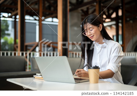Asian woman student with eyeglasses holding pen writing in notebook over laptop on table in cafe. Asian woman student with eyeglasses holding pen writing in notebook over laptop on table in cafe. 134142185