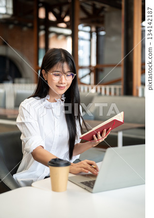 Asian woman student with eyeglasses holding book and looking at laptop as sitting at table in cafe. Asian woman student with eyeglasses holding book and looking at laptop as sitting at table in cafe. 134142187